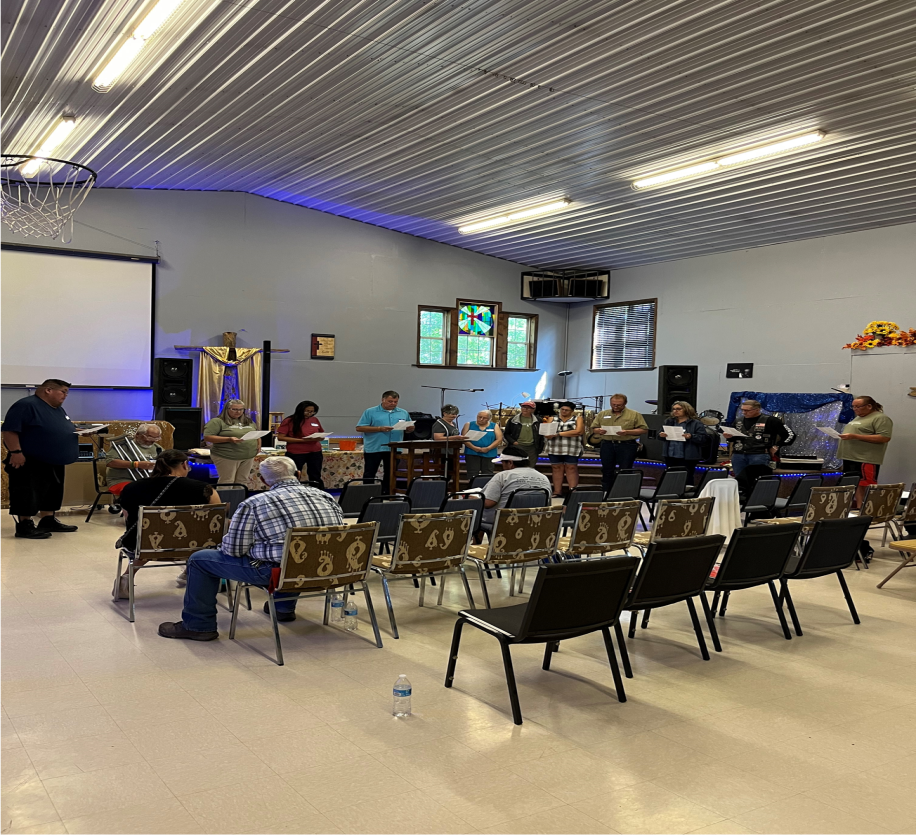 Interior of a multipurpose room with a corrugated metal ceiling and blue accent lighting. Multiple rows of folding chairs face a stage area where approximately 15-20 people are gathered, appearing to participate in a choir or group rehearsal. The room features a basketball hoop on the left wall, a projection screen, windows along the back wall, and religious decorations including what appears to be a purple liturgical cloth. The space has beige walls and flooring, with speakers mounted for sound.